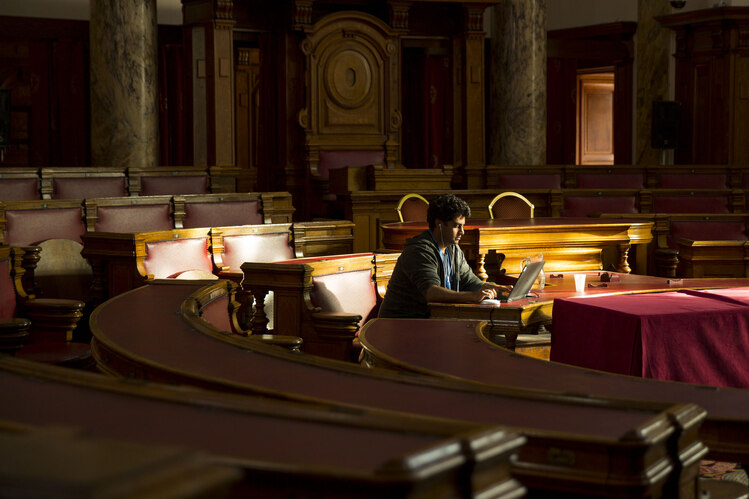 A person working on a laptop in the middle of a council chamber. Most of the room is in shadow.